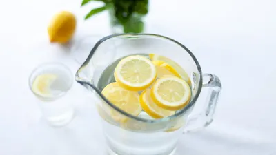 Džbán vody s plátky citronu a sklenicí vody na bílém stole, v pozadí citron a čerstvá máta